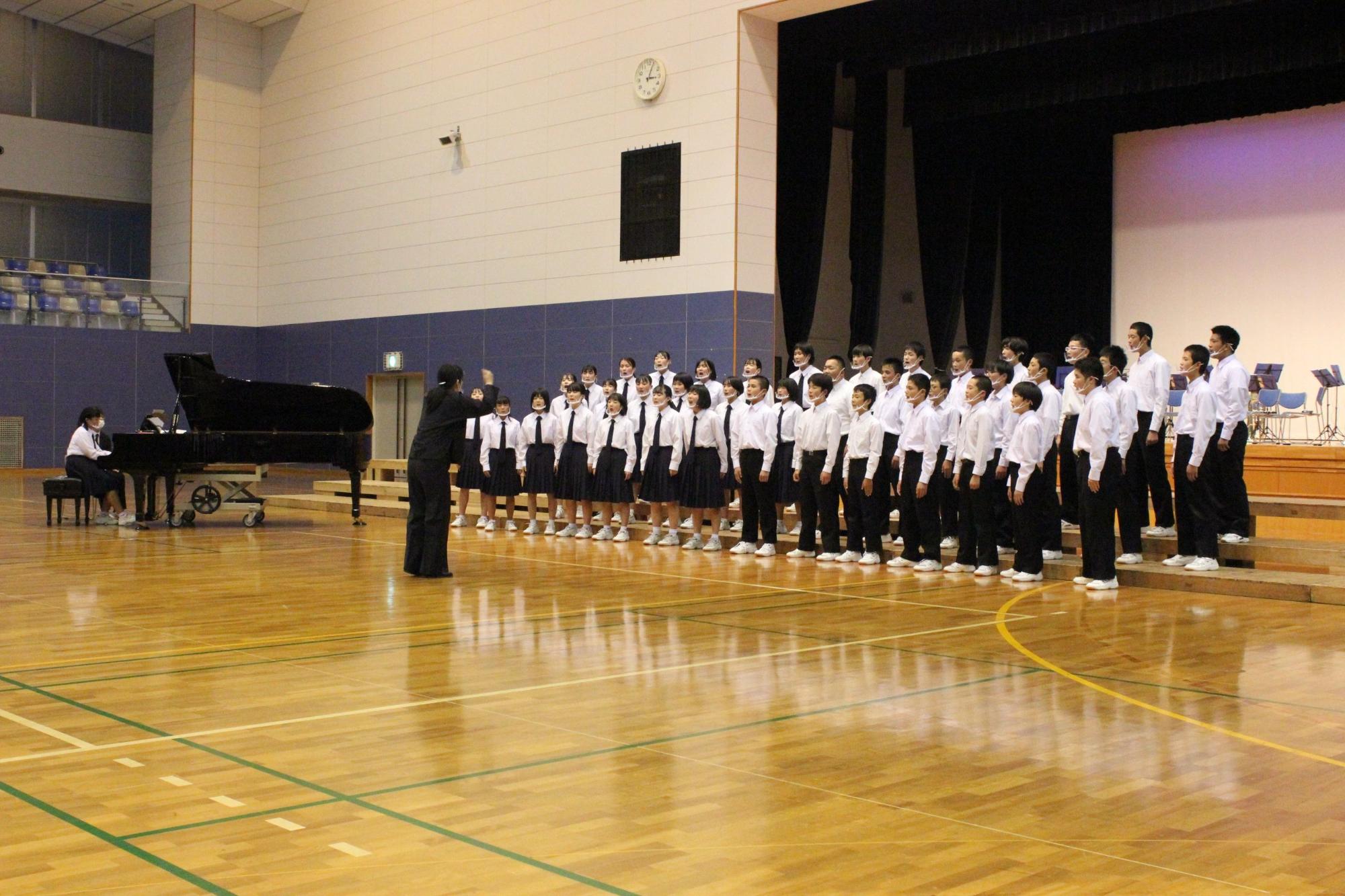 体育館の舞台の前で整列し指揮者に合わせて合唱する数十人の生徒たちの写真