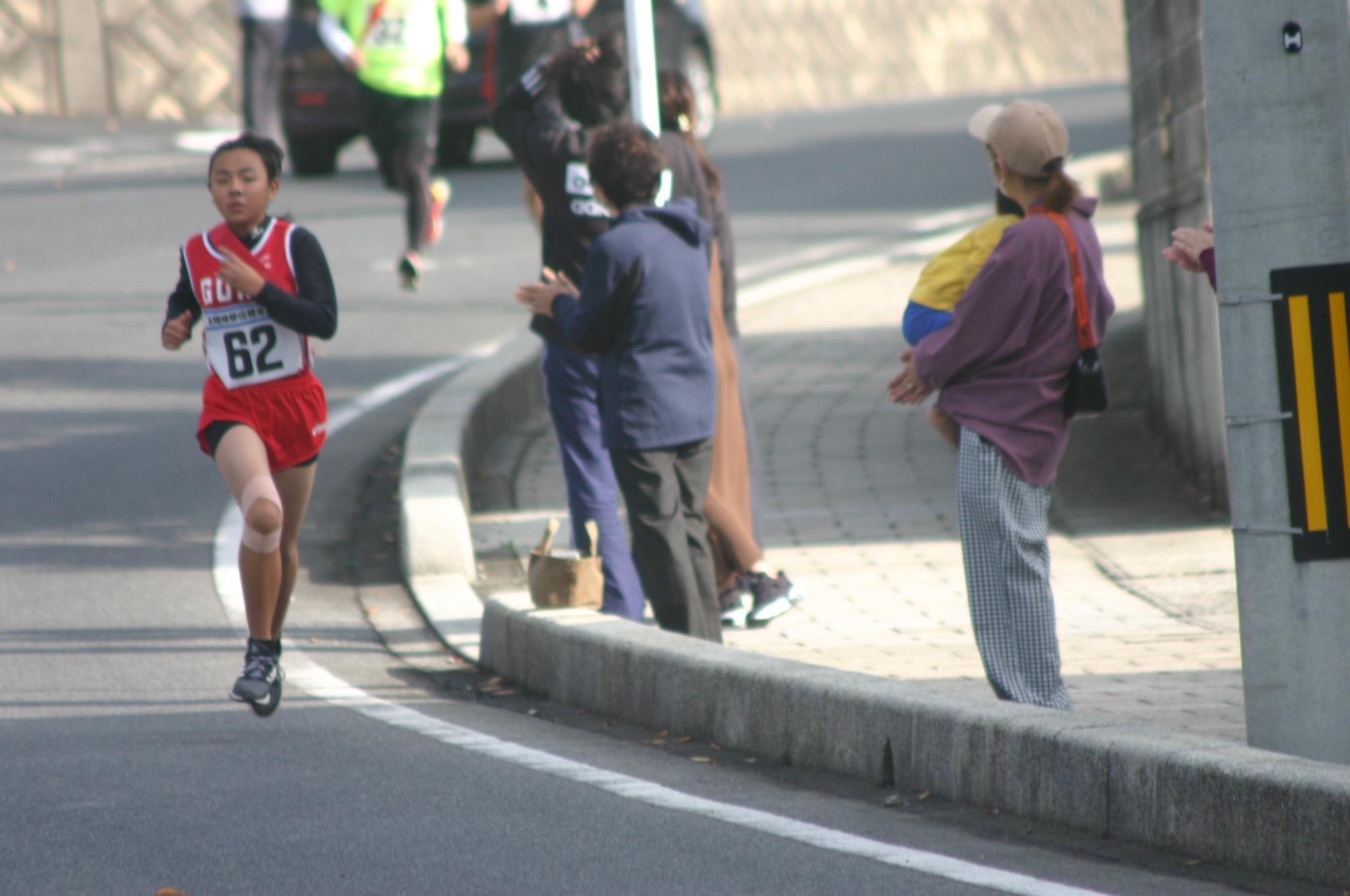 車道の近くを走る赤色のゼッケンを着用した一人の生徒とすぐそばで応援する数人の保護者の写真