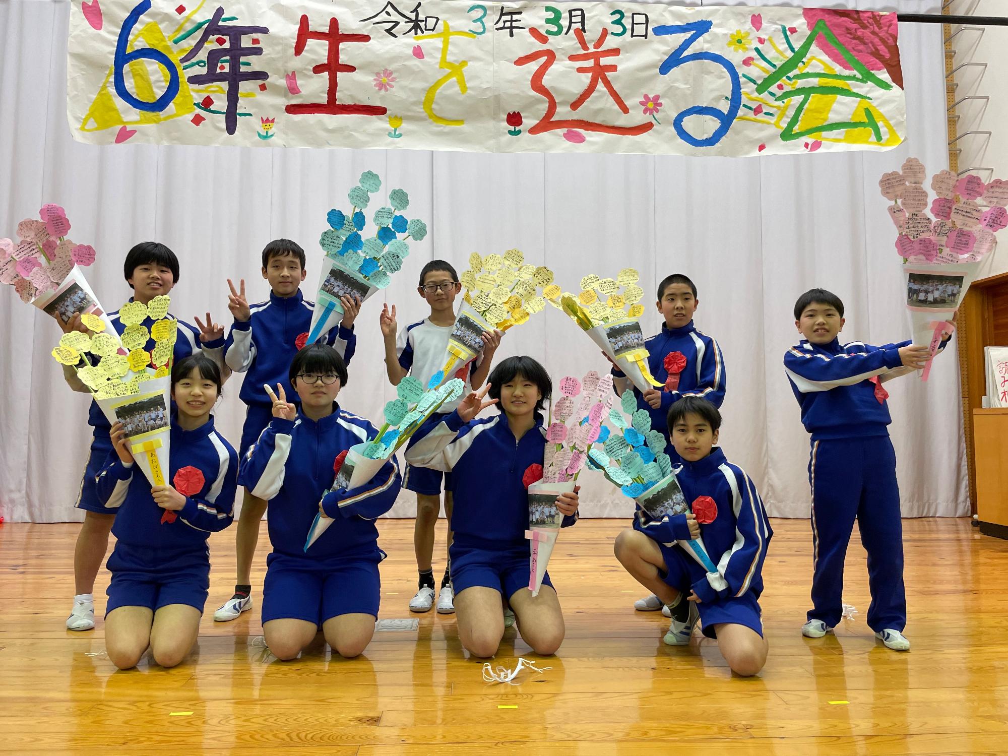 室内で児童たちが「6年生を送る会」と書かれた幕の下で記念撮影を行っている写真