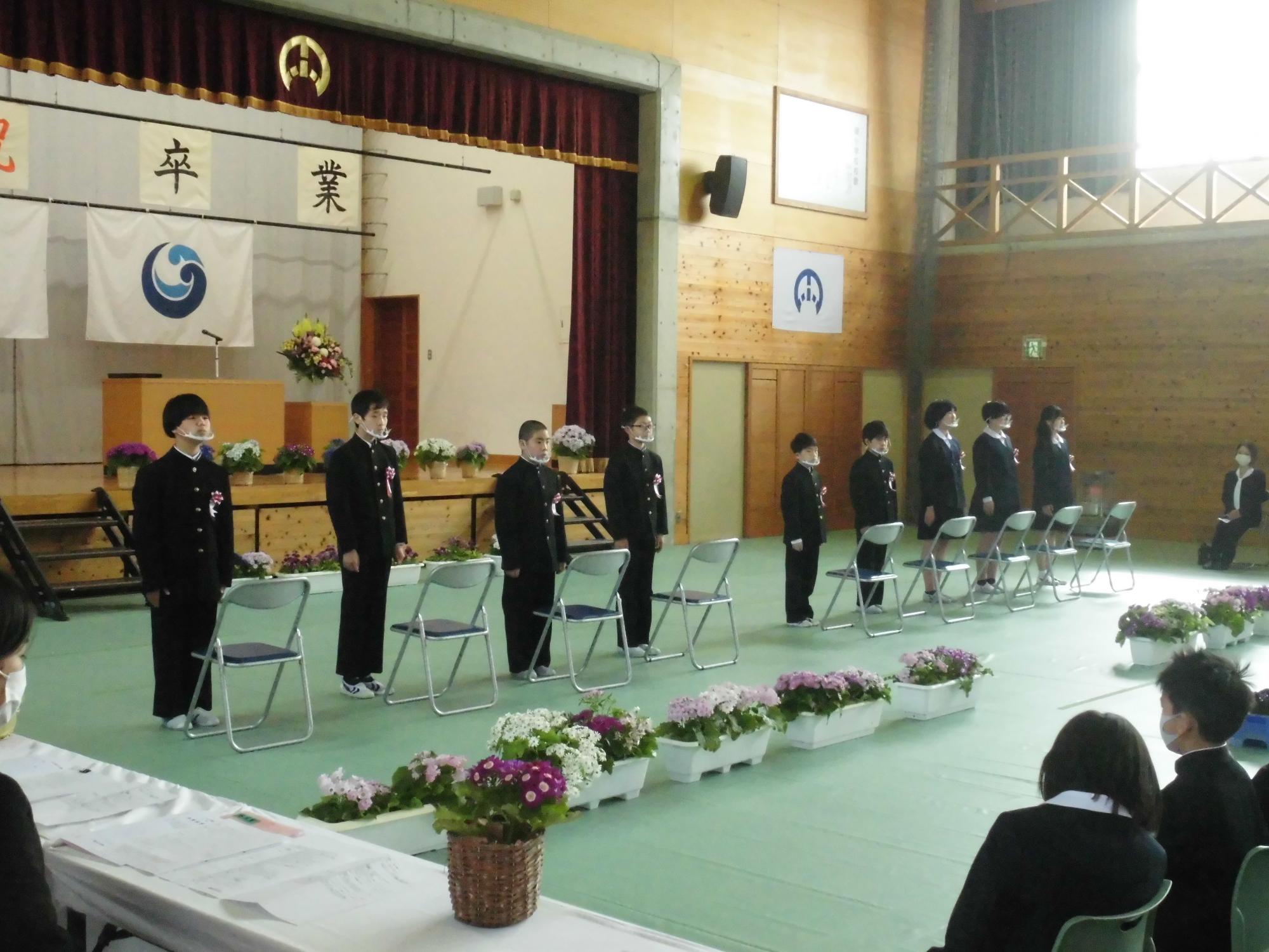 体育館で中学制服を着た児童たちが椅子の前に直立している写真