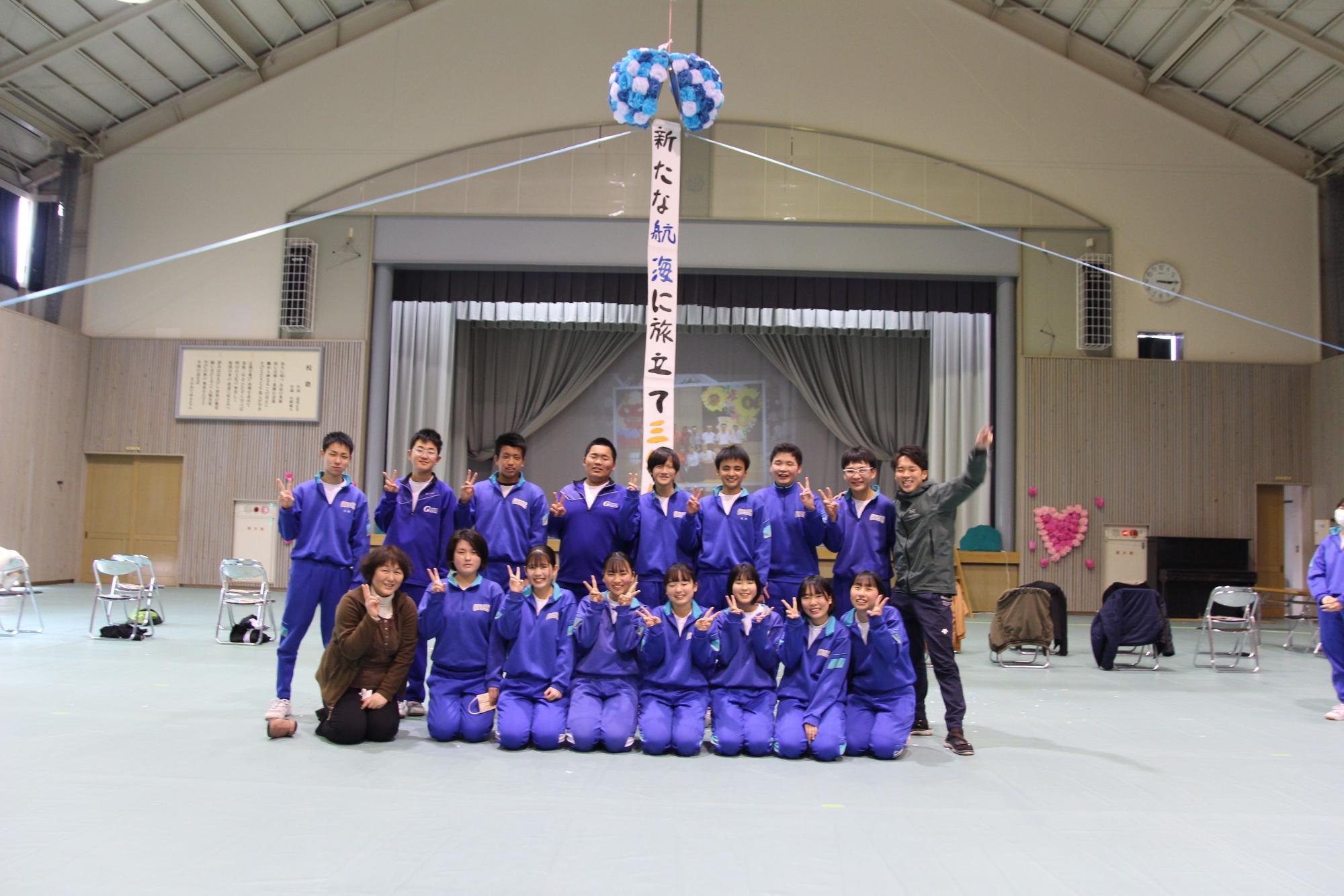 体育館で「新たな航海に旅立て」の垂れ幕の下、集合写真を撮っている写真