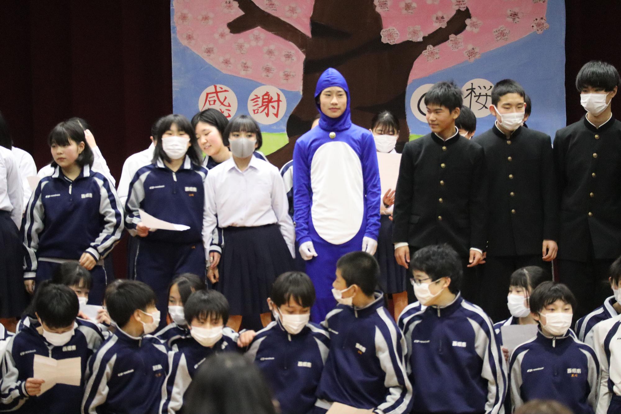 青い着ぐるみを着た男子生徒と、ジャージや制服姿の生徒たちが並んでいる写真
