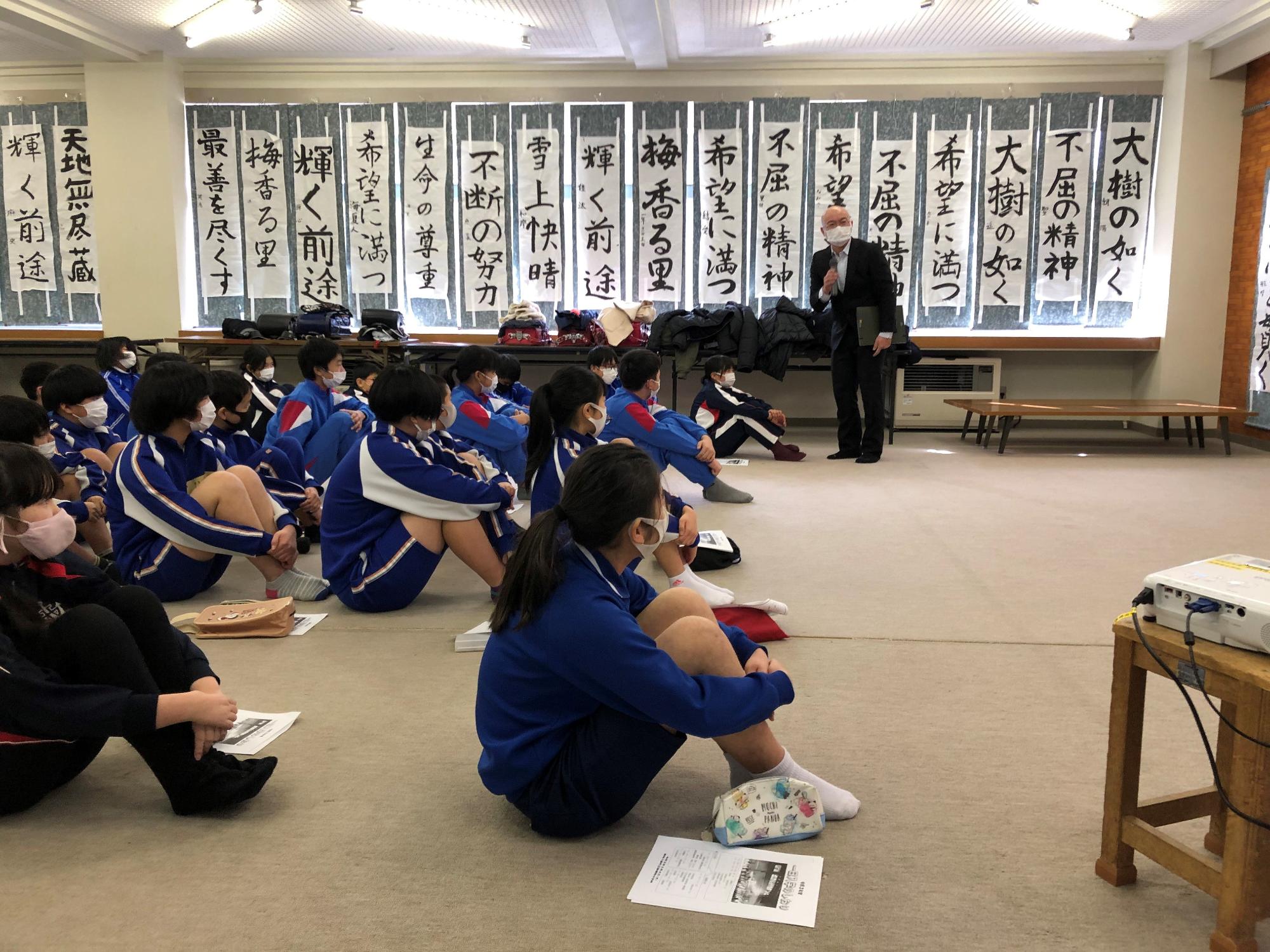 教室でジャージ姿の生徒たちが体育すわりをして先生の話を聞いている写真