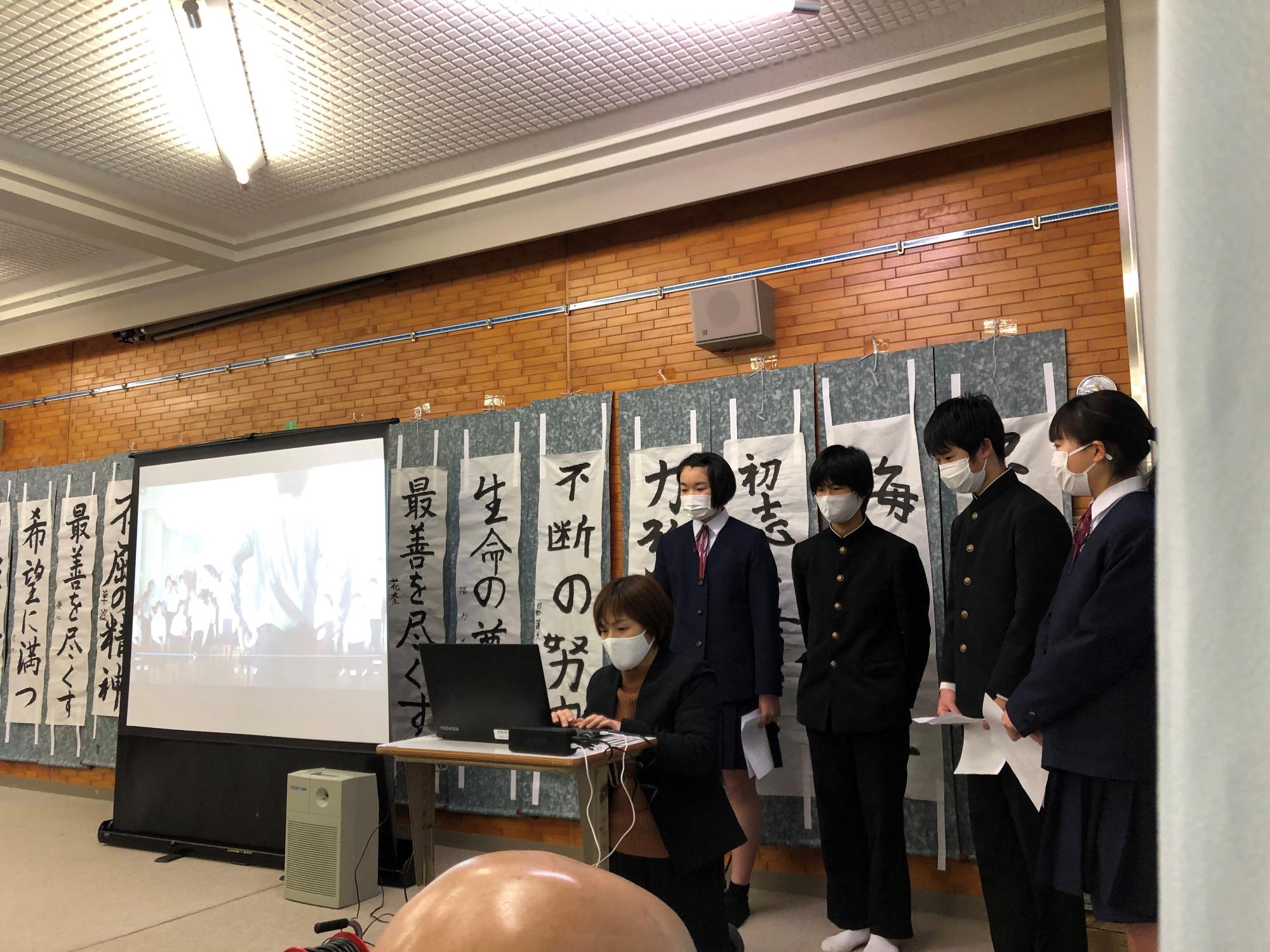 教室に設置されたスクリーンの横に制服姿の生徒と先生が並んでいる様子の写真
