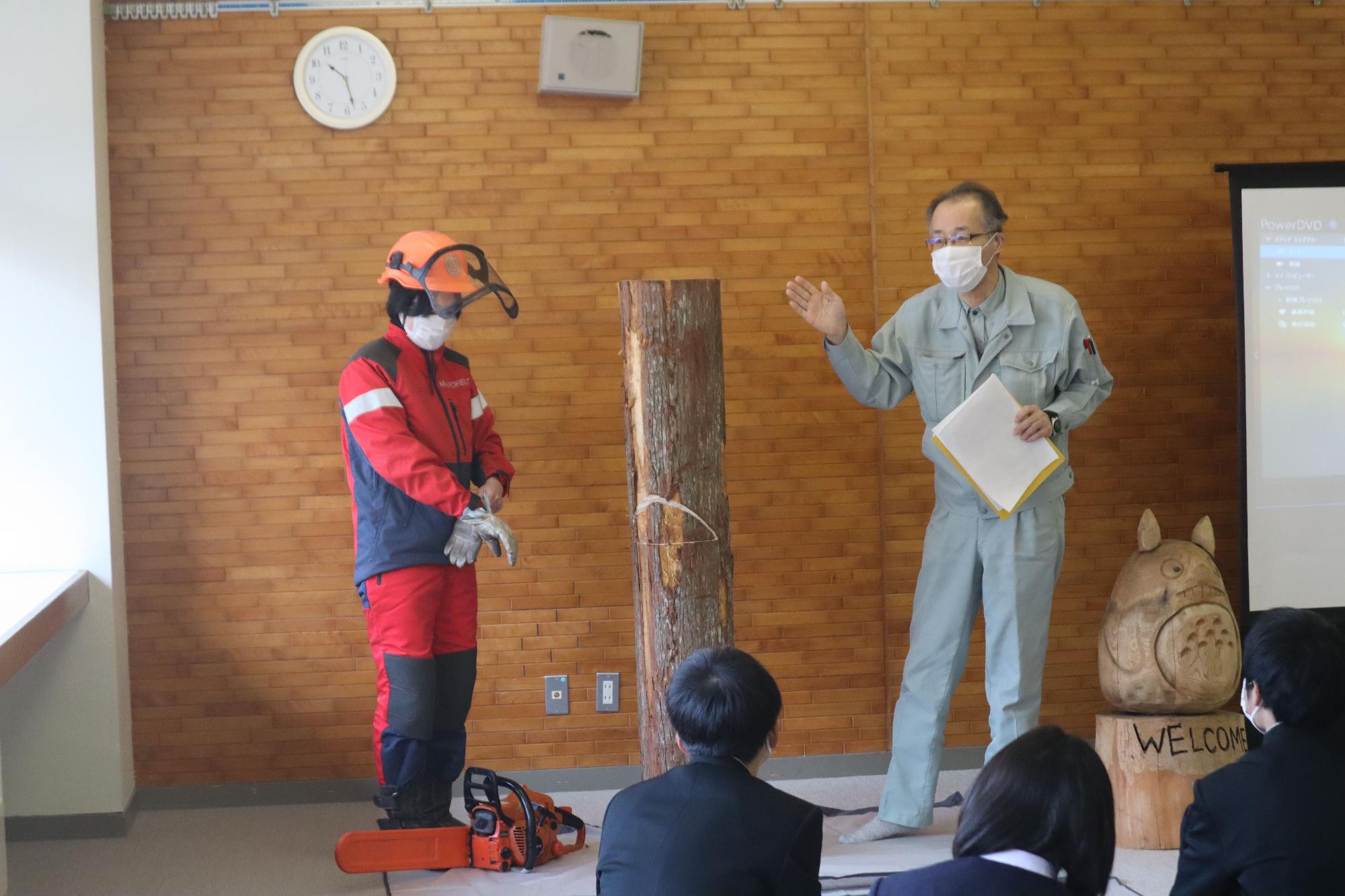 教室の正面に大きな丸太が立てて置かれ、赤い服を着たヘルメットをかぶった男性と、灰色の服を着た男性が立っている様子の写真