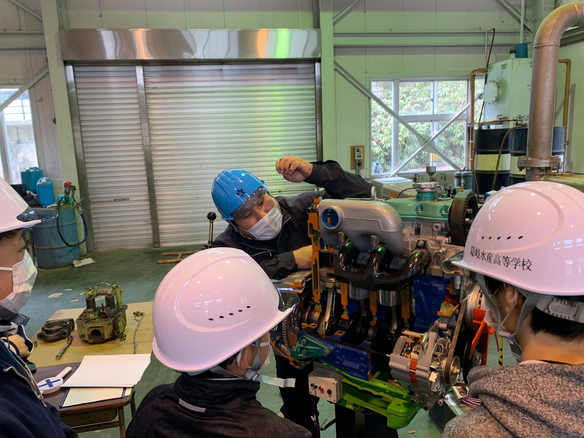 実習場でピンクのヘルメットをかぶった生徒がエンジンの構造を学んでいる写真