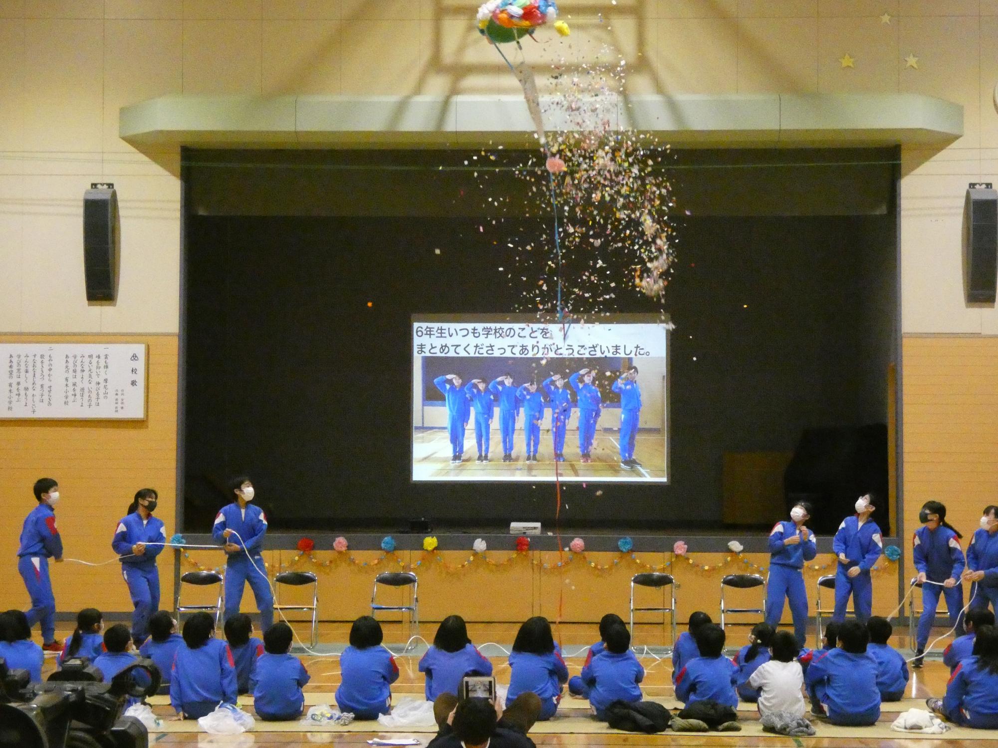 くす玉を割る児童たちと、プロジェクターの感謝のメッセージを見ている数人の児童の写真
