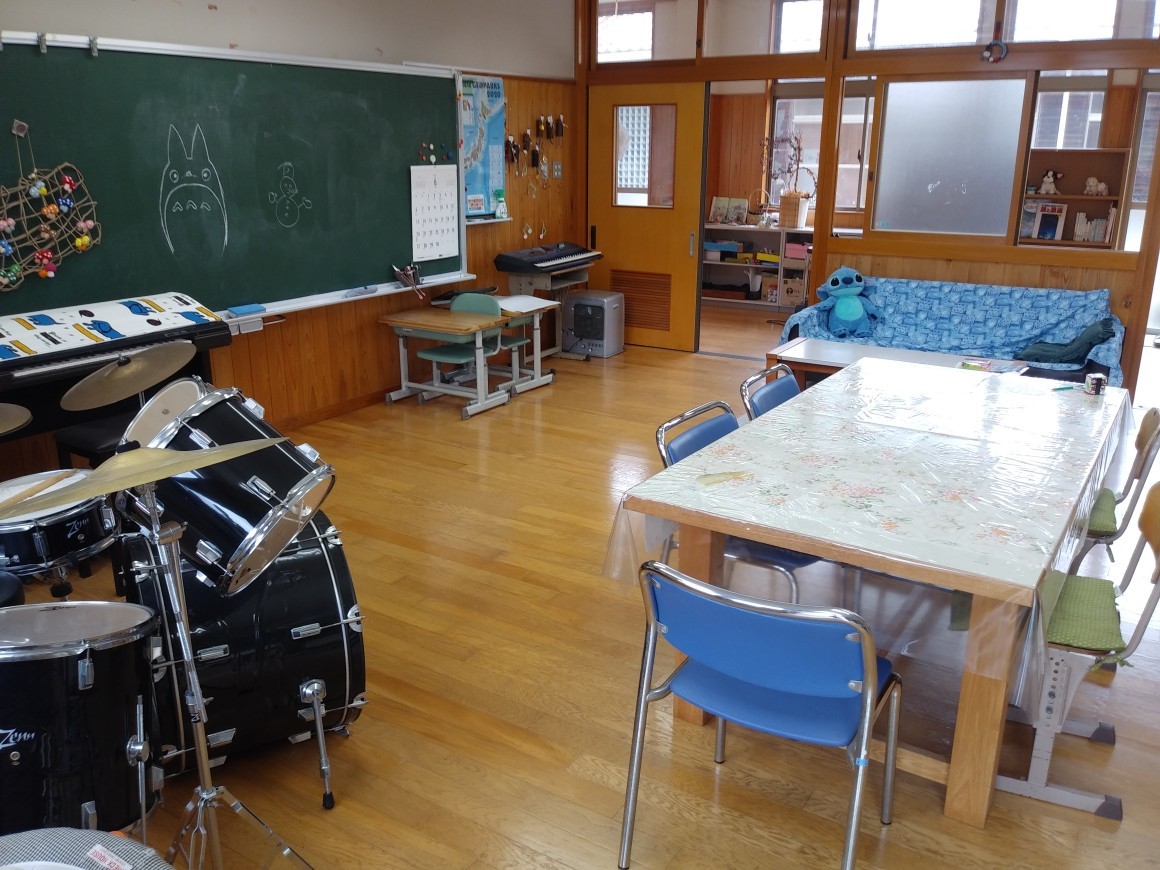 正面に黒板が掛けられテーブルと椅子や楽器のドラムセットなどが置かれた広い室内の様子の写真