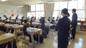 教室の前方で教師が新入生に話しかけている入学初日の教室風景の写真