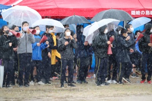 雨の運動場で傘を差した生徒たちが声援を送っている写真