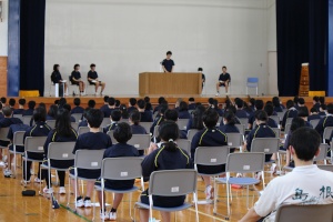 体育館の舞台上で生徒が演壇に立って話をし、他の生徒たちが着席して聞いている写真