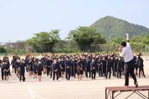 校庭の中央で教師の指示に合わせて生徒たちが体操をしている写真