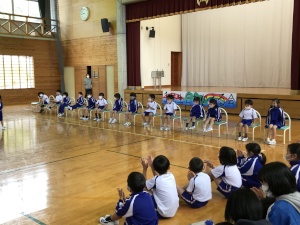体育館で児童たちが椅子に座って並び、児童たちが拍手している写真
