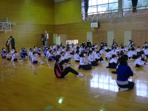 体育館で児童たちや先生が座り、手を使って活動している写真