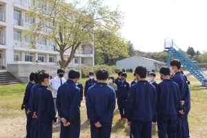 校舎の前のグラウンドで生徒たちが円になって話を聞いている写真