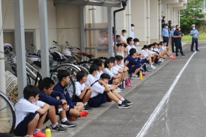 校舎の軒下で講話を聞きながら静かに座る児童たちの写真