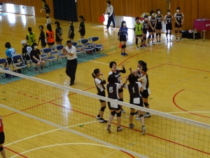 体育館のコートで女子生徒たちがバレーボールの試合中に円陣を組んでいる写真