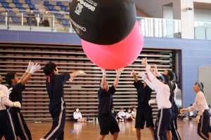 体育館で生徒たちが大きなボールを協力して押し上げている写真