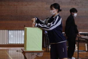 体育館で投票箱に投票用紙を入れている女子生徒の写真