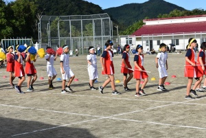 校庭で赤い服と白い服を着た児童たちが隊列を組んで行進している写真