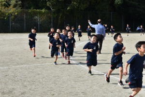 校庭で黒い服を着た児童たちが一列で走っている写真