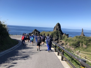 青空の下、児童たちが海沿いの遊歩道を歩いている様子の写真