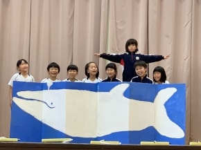 舞台の上でクジラの絵を背景に発表をする児童たちの写真