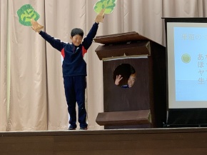 舞台の上で木の模型を持ち発表する児童の写真