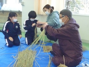室内で地域の人と一緒に藁を編む児童たちの写真