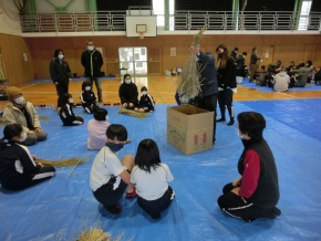 体育館で講師が藁を持ち上げて説明するのを児童たちが見ている写真
