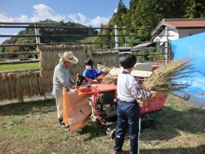 農作業場で児童と地域の大人が脱穀作業をしている写真