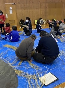 体育館で藁を編む体験をしている児童と地域の人たちの写真