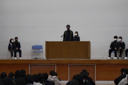 体育館の壇上に立って発表を行っている生徒を正面から撮った写真