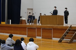 床に座った生徒たちの前で体育館の壇上に立った一人の男子生徒がもう一人の男子生徒と共にマイクで発表を行っている写真