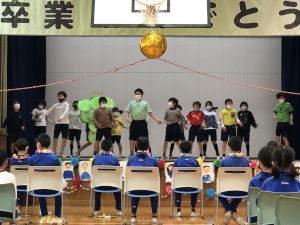 体育館の舞台の上で横一列に並んで出し物を披露する児童たちと、椅子に座ってそれを観覧している児童たちの様子の写真