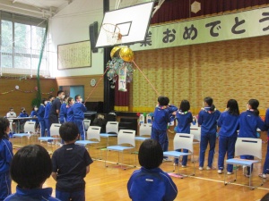 体育館の中でバスケットボールのゴールリングから吊された金色のくす玉を、児童たちが左右から紐を引いて割っている瞬間を捉えた写真