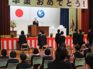 体育館の壇上の演壇で話すスーツを着た男性と、それを椅子から起立して聞き入る卒業生児童たちの様子の写真