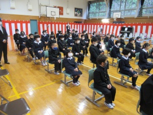 体育館の中の壁にぐるりと張られた紅白の横幕の前で、並べられた椅子に着座している児童たちの写真