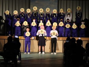 体育館の舞台で生徒たちが座っている三年生に感謝のメッセージを掲げている写真