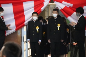 入学式で黄色い花を付けた新入生が入場する様子の写真