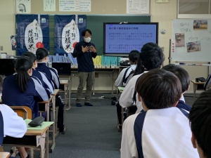 教室で正面に置かれたモニターに映し出された資料を見ながら、講師のを聞く生徒たちの写真