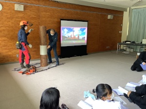 教室正面のモニターの前で太い木の模型で説明を行う2人の講師の写真