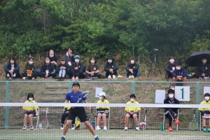 テニスの試合を観戦する生徒たちの応援を受けながら試合をする生徒の写真