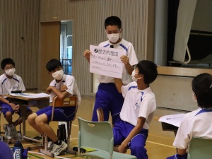 体育館で発表する男子生徒と椅子に座り話を聞いている生徒たちの写真