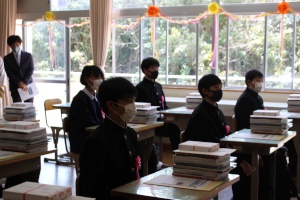 教室で新入生が教科書が積まれた机に向かって着席している写真