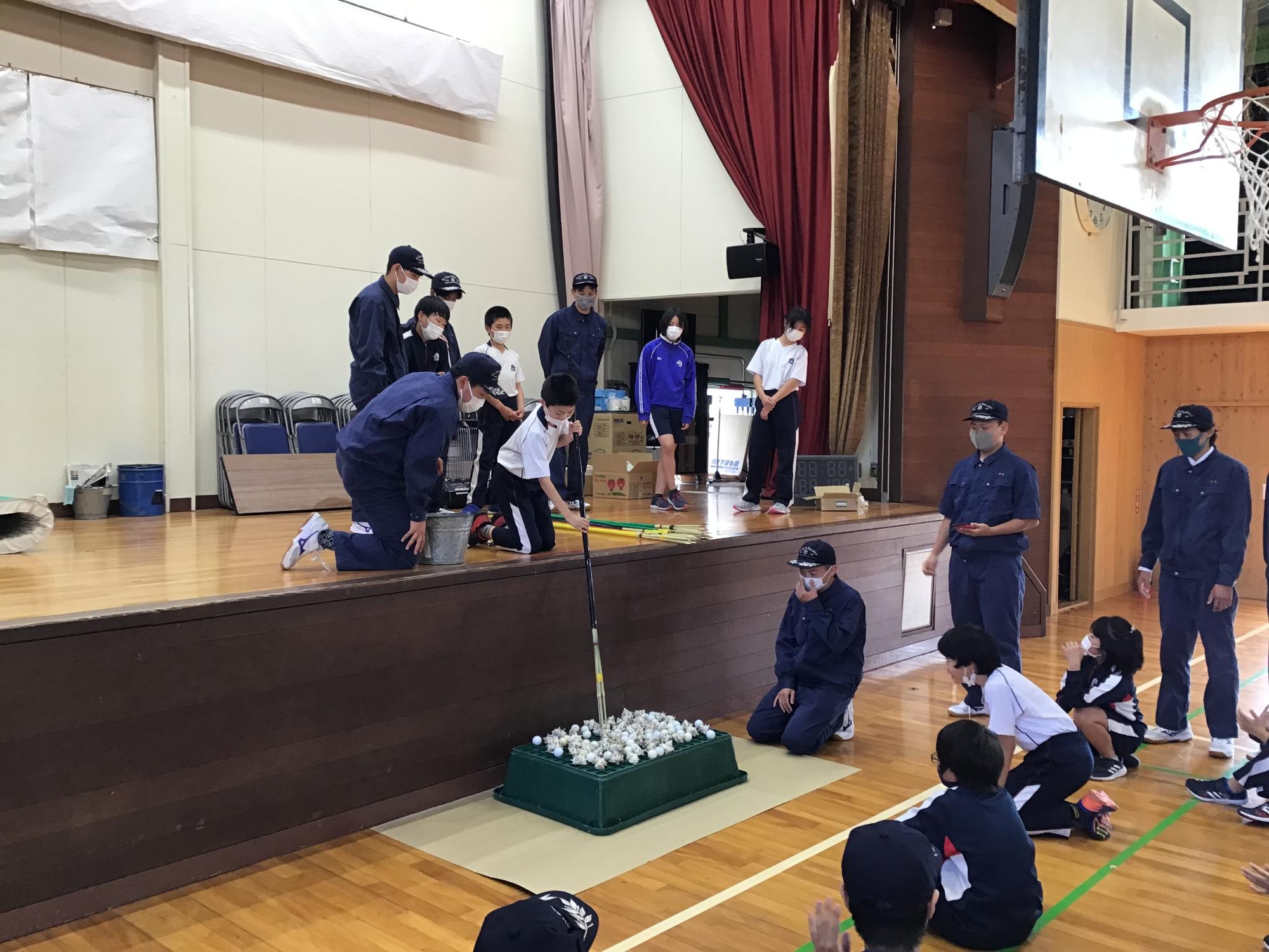 体育館の舞台上で高校生が小学生に伝統の漁の説明をしている写真