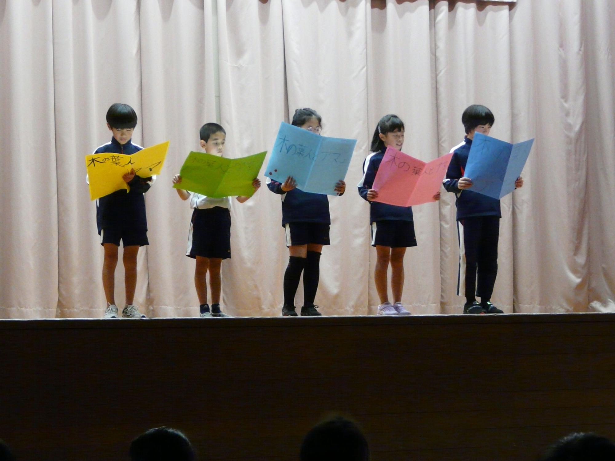 舞台上で黄色や水色などカラフルな色の大きな紙を掲げて発表する児童たちの写真