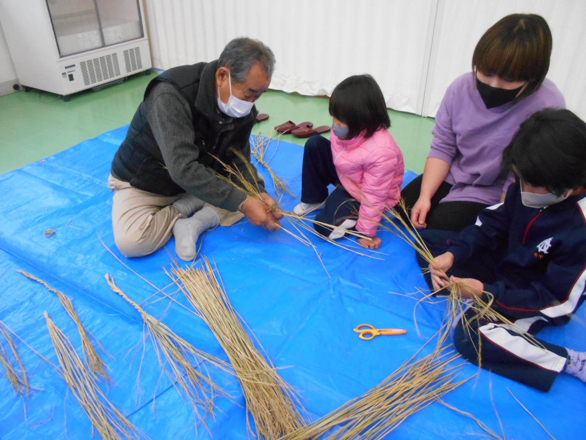 室内で大人と子供が藁を使ってしめ縄作りをしている写真