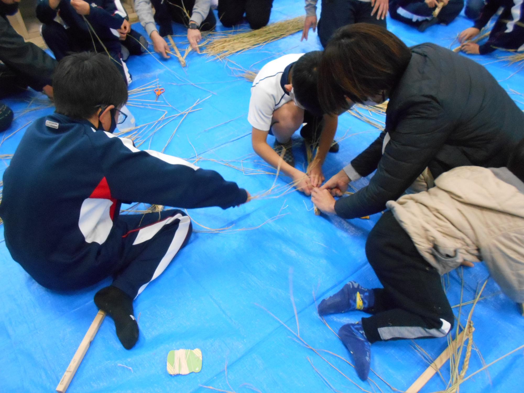 室内で児童と大人が協力して藁を編んでいる写真