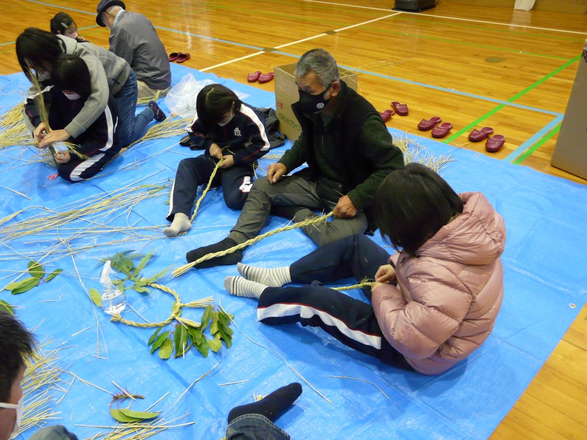 体育館で児童と地域の人が一緒にしめ縄作りをしている写真